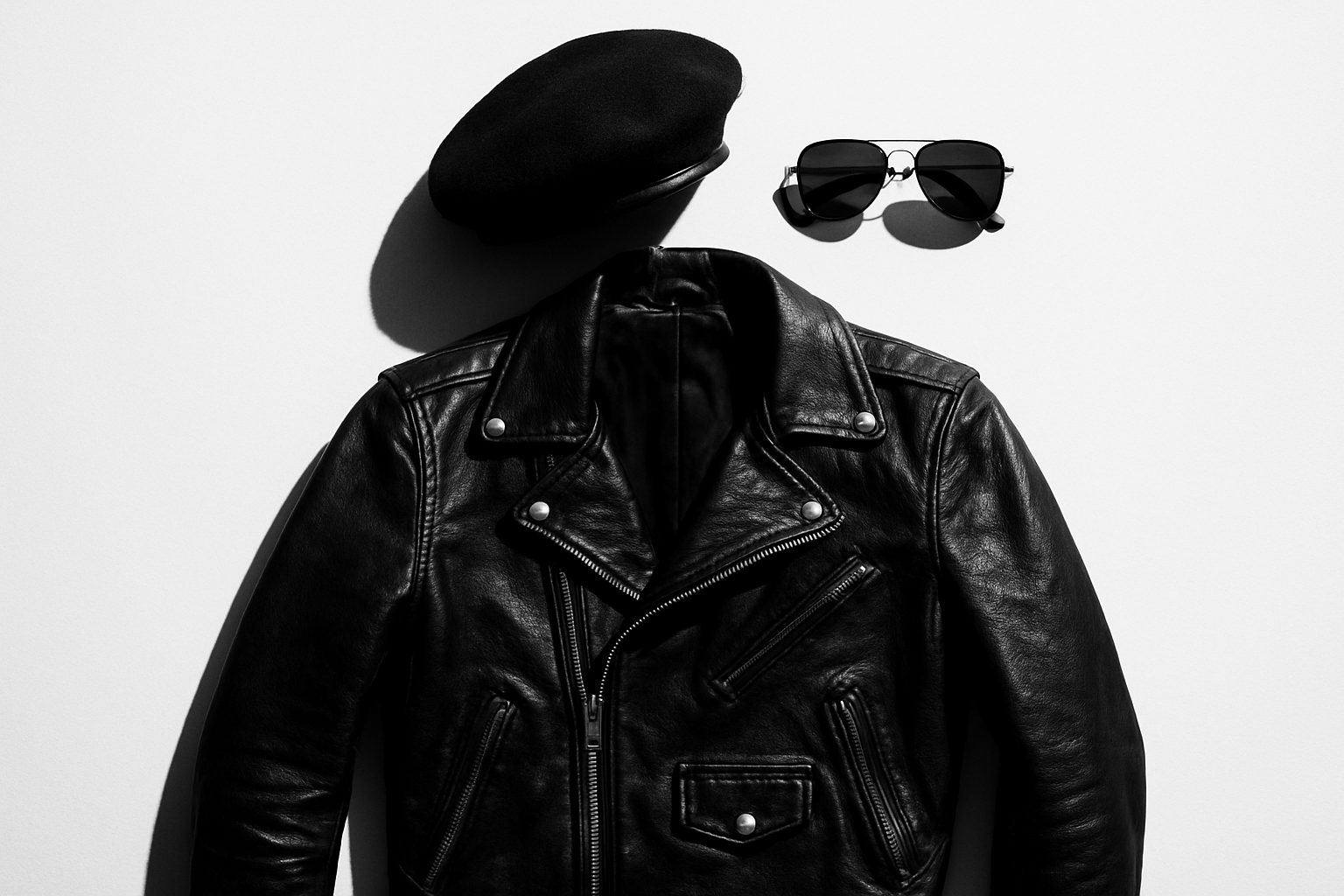 A flat-lay arrangement of a black leather jacket, black beret, and dark aviator sunglasses on a white background representing the iconic Black Panther Party aesthetic